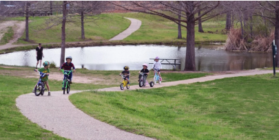 Children riding bikes along a paved path beside a pond at Northwest District Park in Allandale, with grass and trees along the trail